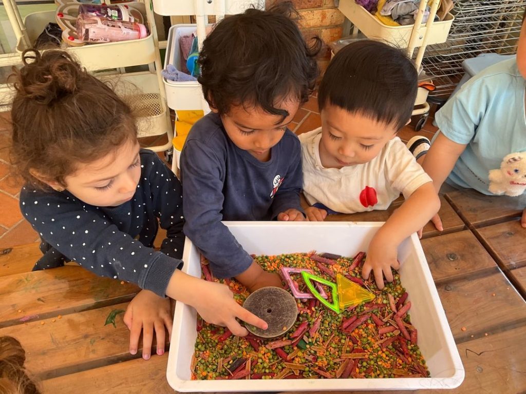 3 children playing with shapes in a sensory tray.