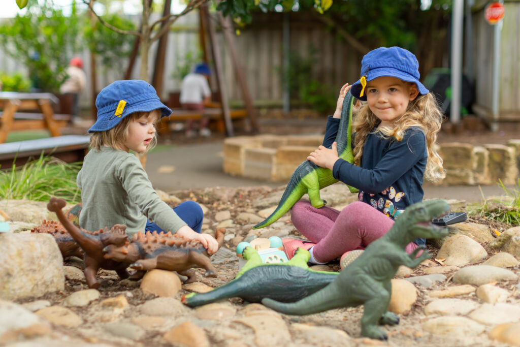 Two children playing outside with toy dinosaurs. They are both wearing Care for Kindies hats.